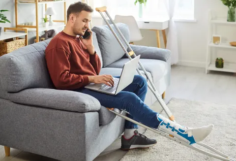 Junger Mann mit Gipsbein und Krücken sitzt telefonierend vor einem Laptop auf dem Sofa.