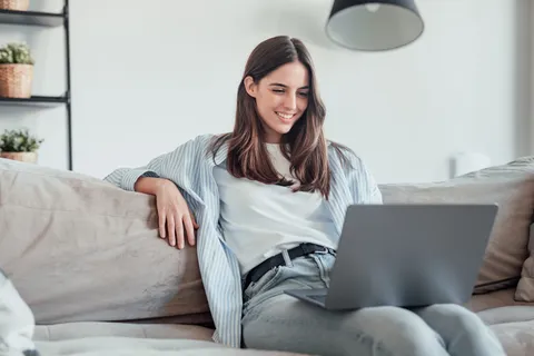 Junge Frau sitzt entspannt auf der Couch mit Laptop auf den Beinen