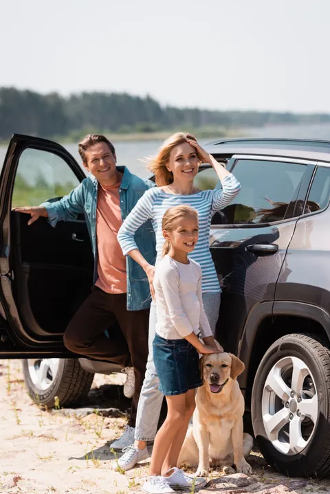 Frau und Mann stehen mit ihrer Tochter und Hund vor einem Auto am See.