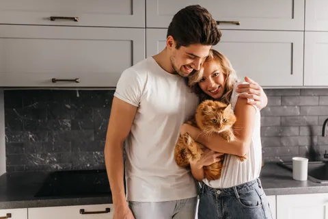 Eine junge Frau hält eine rote Katze im Arm und wird von einem jungen Mann umarmt.