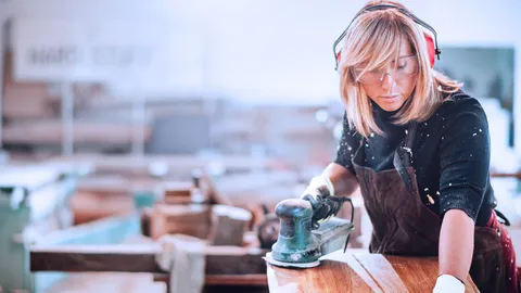 Eine Frau in Arbeitskleidung schleift in einer Werkstatt Holz