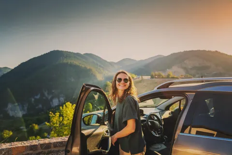 Eine junge Frau steht vor ihrem Auto und blickt über die geöffnete Fahrzeugtür auf die Berge im Hintergrund