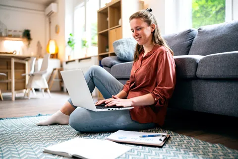 Junge Frau sitzt auf dem Boden, mit Couch im Rücken und Laptop auf den Beinen, ein aufgeschlagenes Buch und ein Notizblock daneben