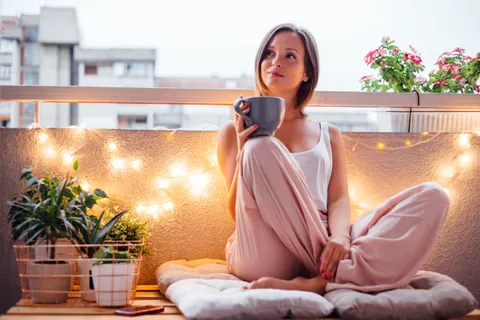 Junge Frau sitzt gemütlich mit einer Tasse Kaffee auf ihrem Balkon im Schneidersitz