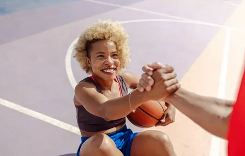 einer auf dem Boden sitzenden Frau mit Basketball unter dem Arm wird beim Aufstehen geholfen