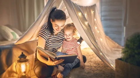 Junge Mutter sitzt mit kleiner Tochter in einem Zelt in der Wohnung und liest ein Buch vor